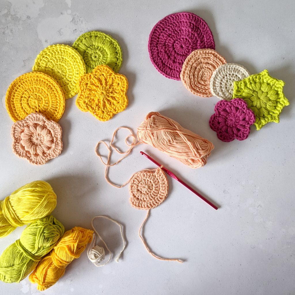 Ten finished crochet face scrubs in round and flower shapes made in yellow, green, peach and pink yarn. There is also a face scrub in progress