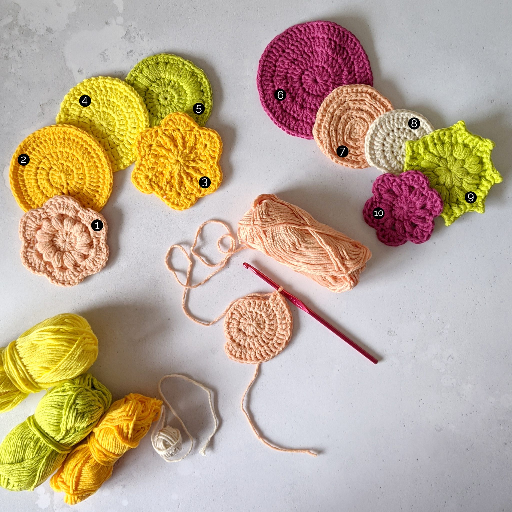 Ten finished crochet face scrubs in round and flower shapes made in yellow, green, peach and pink yarn. There is also a face scrub in progress. Marked up with numbers 1- 10