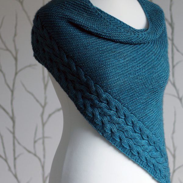 A blue garter stitch shawl with a rippling cable pattern down one side wrapped around a mannequin