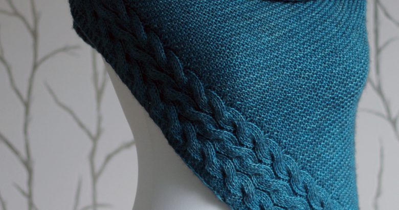 A blue garter stitch shawl with a rippling cable pattern down one side wrapped around a mannequin