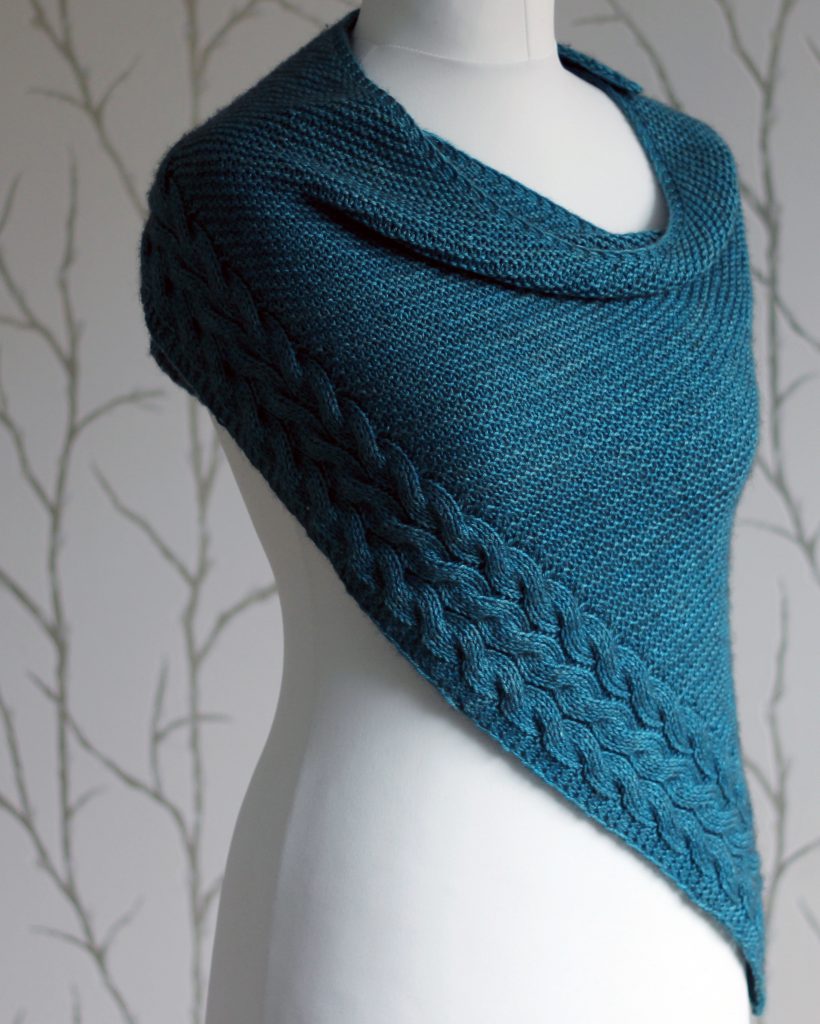 A blue garter stitch shawl with a rippling cable pattern down one side wrapped around a mannequin