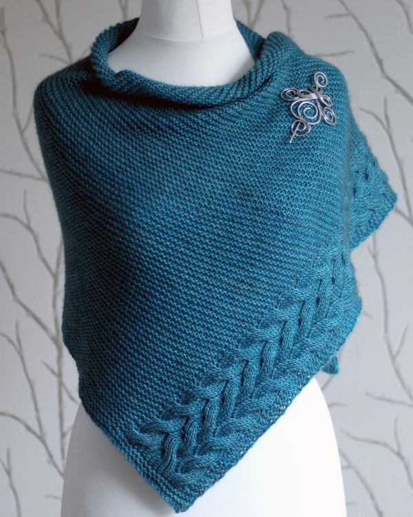 A blue garter stitch shawl with a rippling cable pattern down one side wrapped around a mannequin
