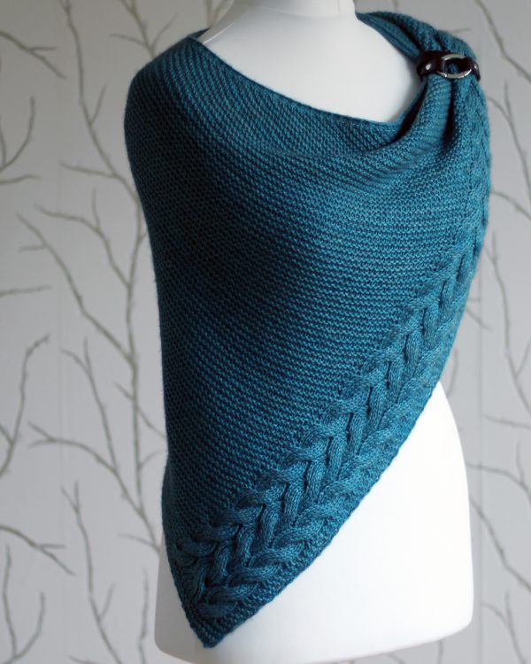 A blue garter stitch shawl with a rippling cable pattern down one side wrapped around a mannequin