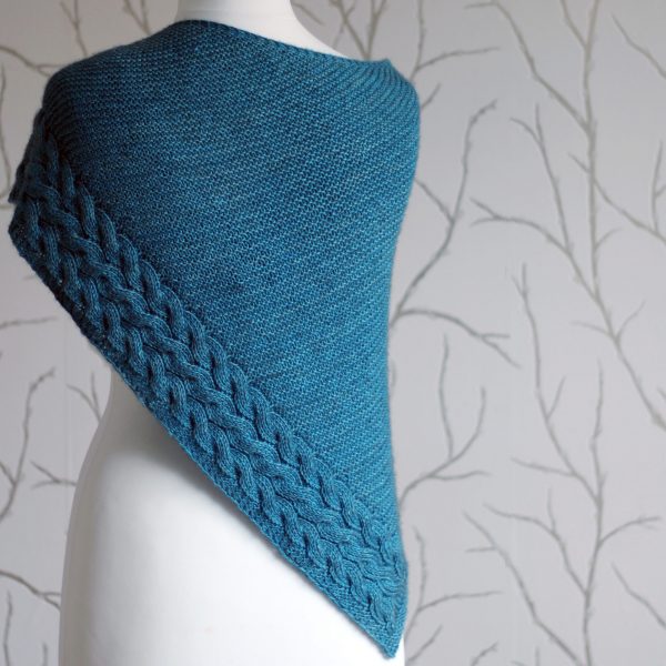 A blue garter stitch shawl with a rippling cable pattern down one side wrapped around a mannequin