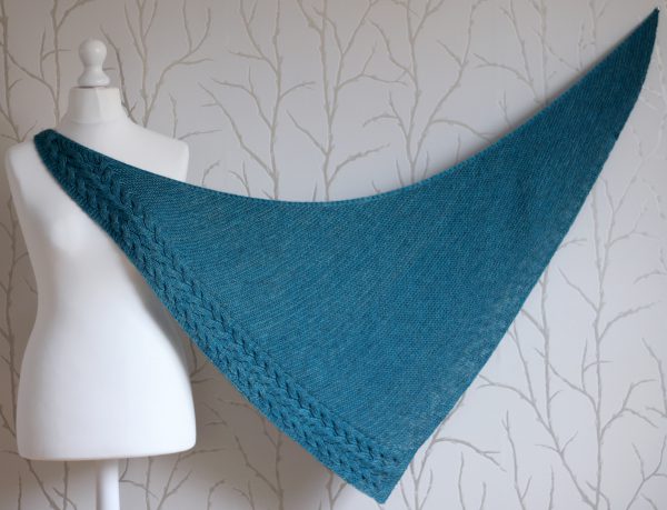 A blue garter stitch shawl with a rippling cable pattern down one side stretched to show the shape