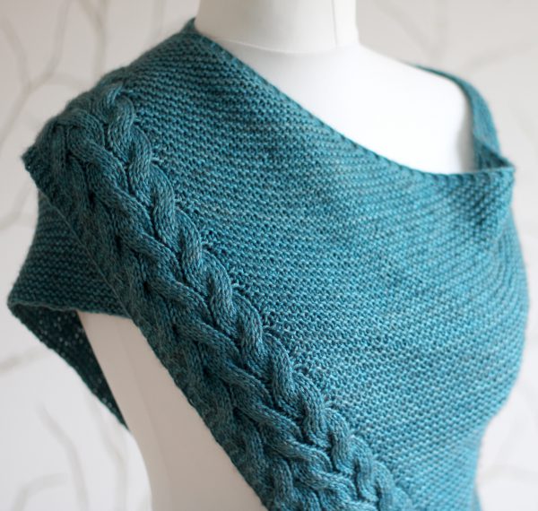 A blue garter stitch shawl with a rippling cable pattern down one side wrapped around a mannequin