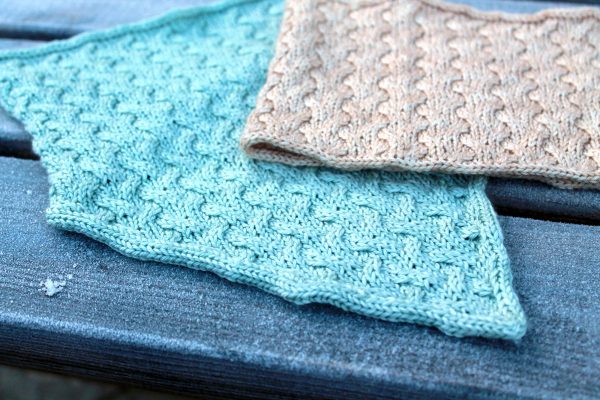 A close up of two cowls with cables and i-cord edging. one has a straight bottom edge and one has a pointed edge