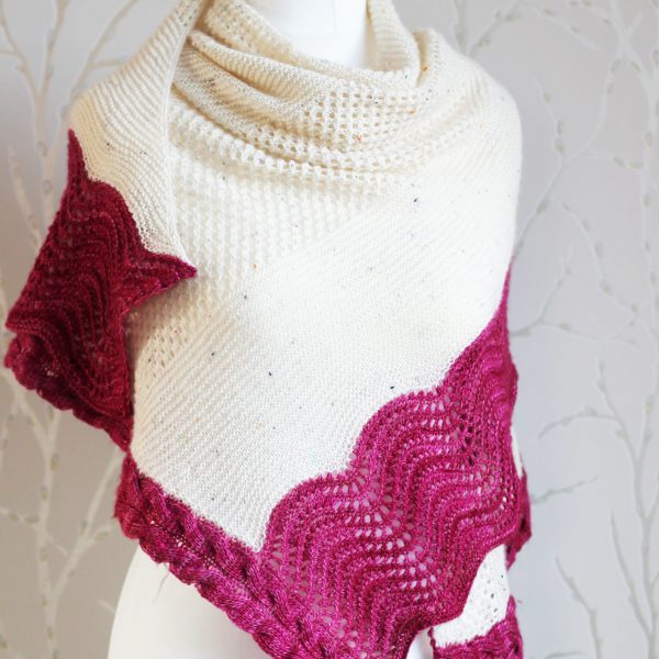 A white triangular shawl with lace and garter stitch stripes and a pink intarsia cable down the side and wavey pink border