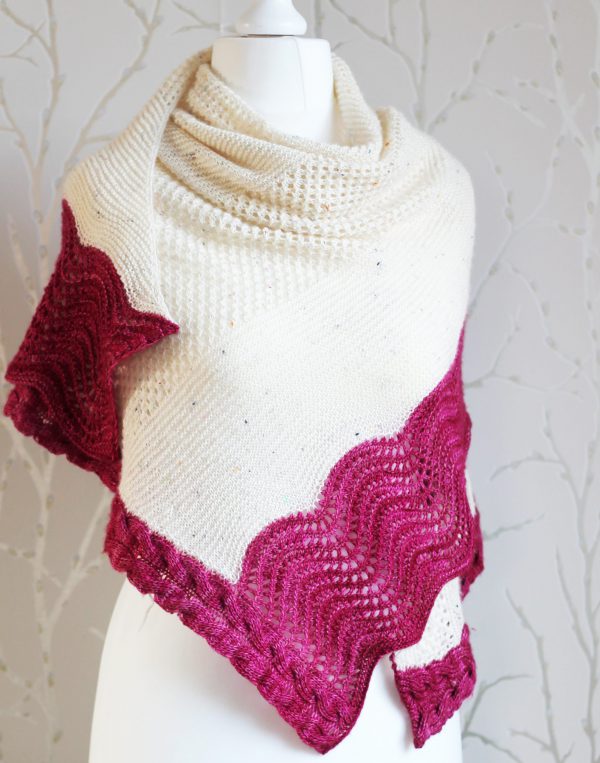 A white triangular shawl with lace and garter stitch stripes and a pink intarsia cable down the side and wavey pink border