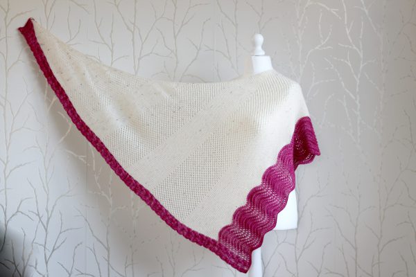 A white triangular shawl with lace and garter stitch stripes and a pink intarsia cable down the side and wavey pink border