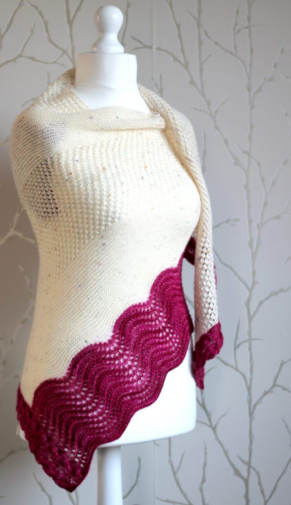 A white triangular shawl with lace and garter stitch stripes and a pink intarsia cable down the side and wavey pink border