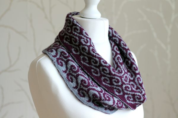 A colourwork cowl with purple waves against a grey background on the outside and grey swirls against a purple background on the inside which has been folded down