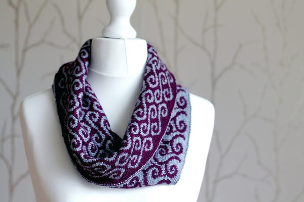 A colourwork cowl with purple waves against a grey background on the outside and grey swirls against a purple background on the inside which has been folded down