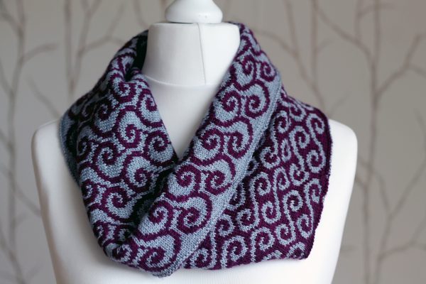 A colourwork cowl with grey swirls against a purple background on the outside and purple waves against a grey background on the inside which has been folded down