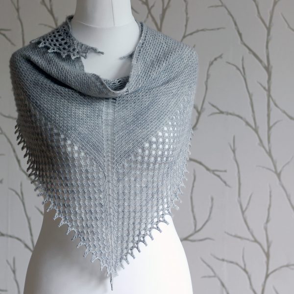 A grey shawlette with a textured body and wide lace border on a white mannequin.