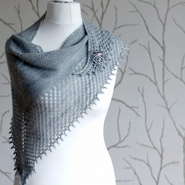 A grey shawlette with a textured body and wide lace border on a white mannequin.