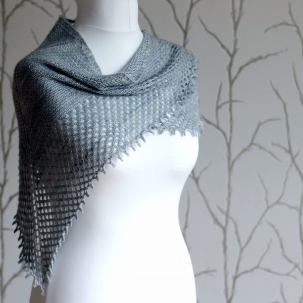 A grey shawlette with a textured body and wide lace border on a white mannequin.