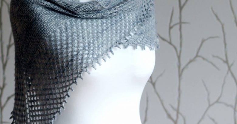 A grey shawlette with a textured body and wide lace border on a white mannequin.