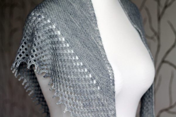 A grey shawlette with a textured body and wide lace border on a white mannequin.