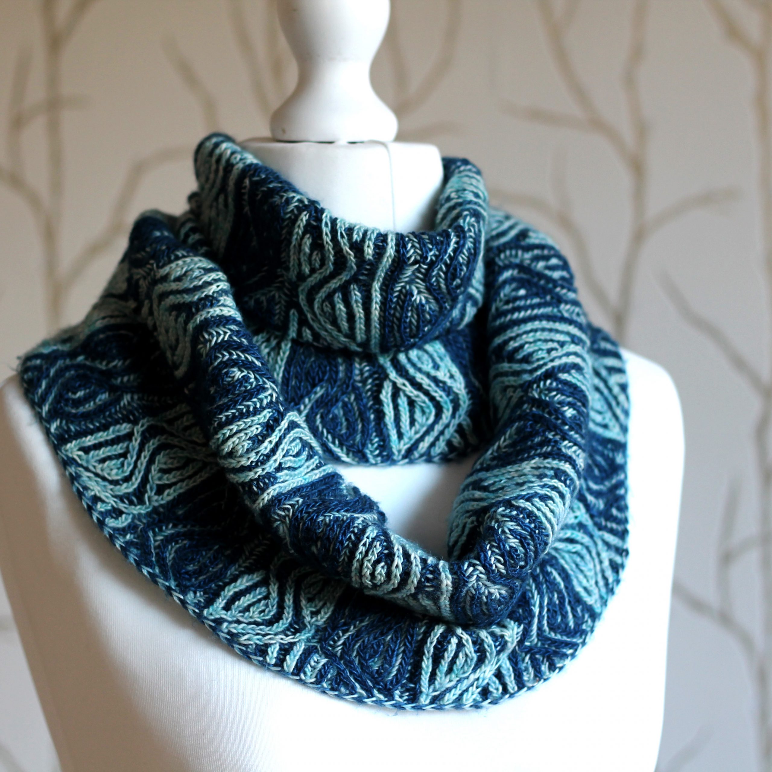 A mannequin displaying a long brioche cowl with dark and light blue vertical stripes and a faux cable pattern wrapped twice around the neck