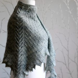 A grey shawl with a cabled spine, a garter stitch body with slipped stitch detail and chevron detail on the edge.