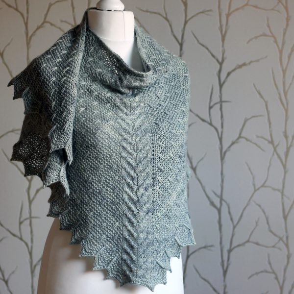 A grey shawl with a cabled spine, a garter stitch body with slipped stitch detail and chevron detail on the edge.