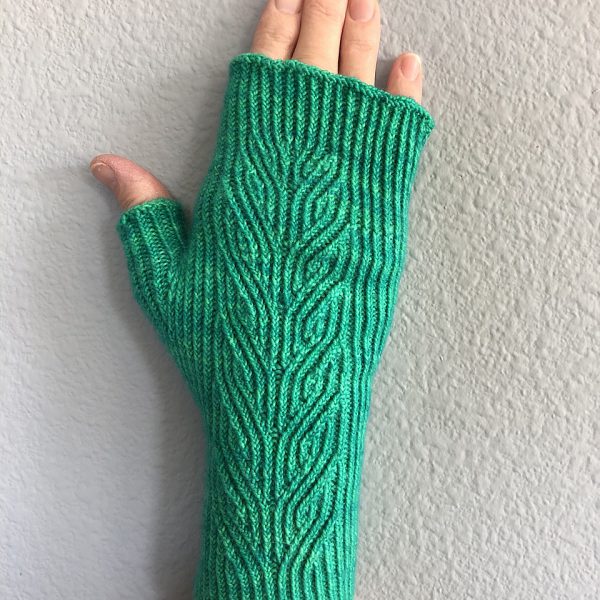 A green fingerless mitt with twisted rib and a faux cable pattern, against a white wall