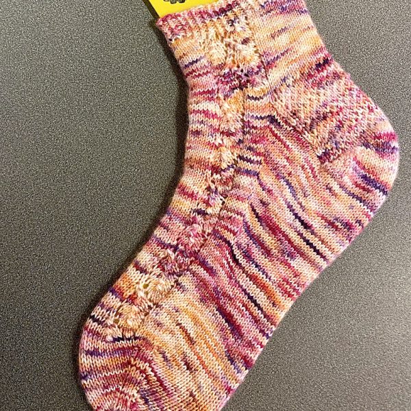 A sock with a lace leaf pattern up the outside of the foot knitted in variegated pink yarn