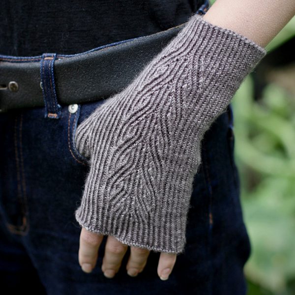 A twisted rib fingerless mitt with a sweeping faux cable pattern up the back of the hand in grey yarn, with the thumb caught in the pocket of the wearer