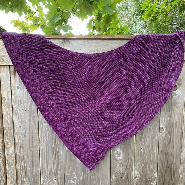 An asymmetrical garter stitch shawl with a reversible cable pattern down one edge hanging over a fence