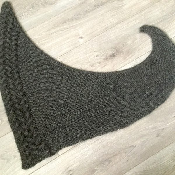 A dark grey asymmetrical triangular shawl with a cable down one side laid on the floor