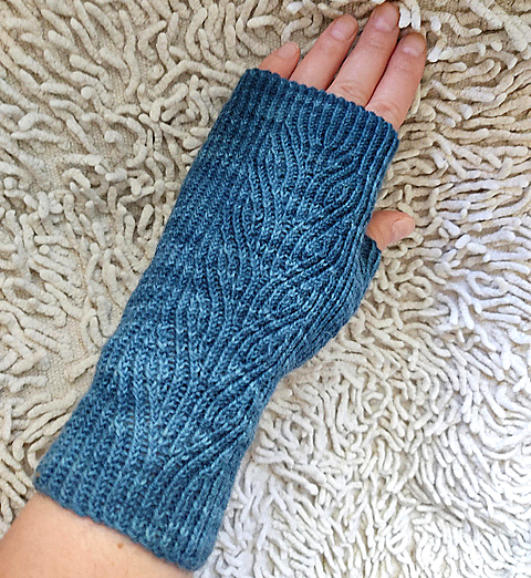 A blue fingerless mitt with twisted rib and a faux cable pattern, against a textured white rug