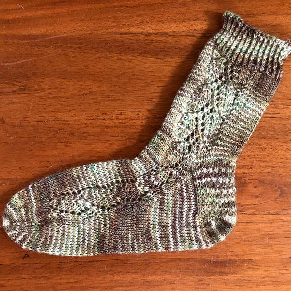 A sock with a lace leaf pattern up the outside of the foot knitted in variegated brown and white yarn