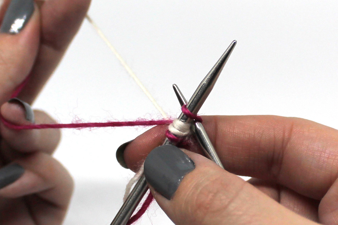 The right hand needle has been inserted between the first two stitches on the left hand needle. The white yarn is being lifted from under the pink yarn in preparation for the stitch.