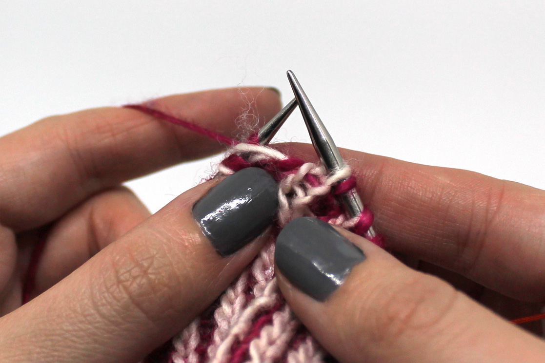 The white yarn has been laid over the first stitch on the left hand needle and is held in place