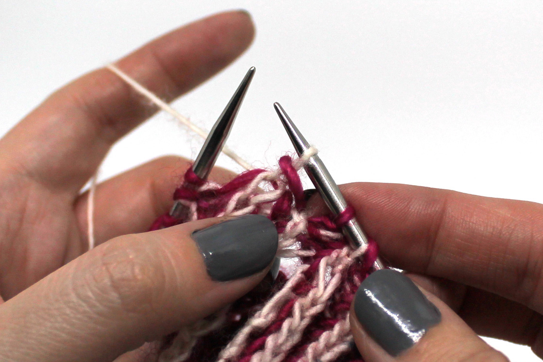 A close up of the bind off in progress. A pink stitch has been lifted over the white stitch and off the needle