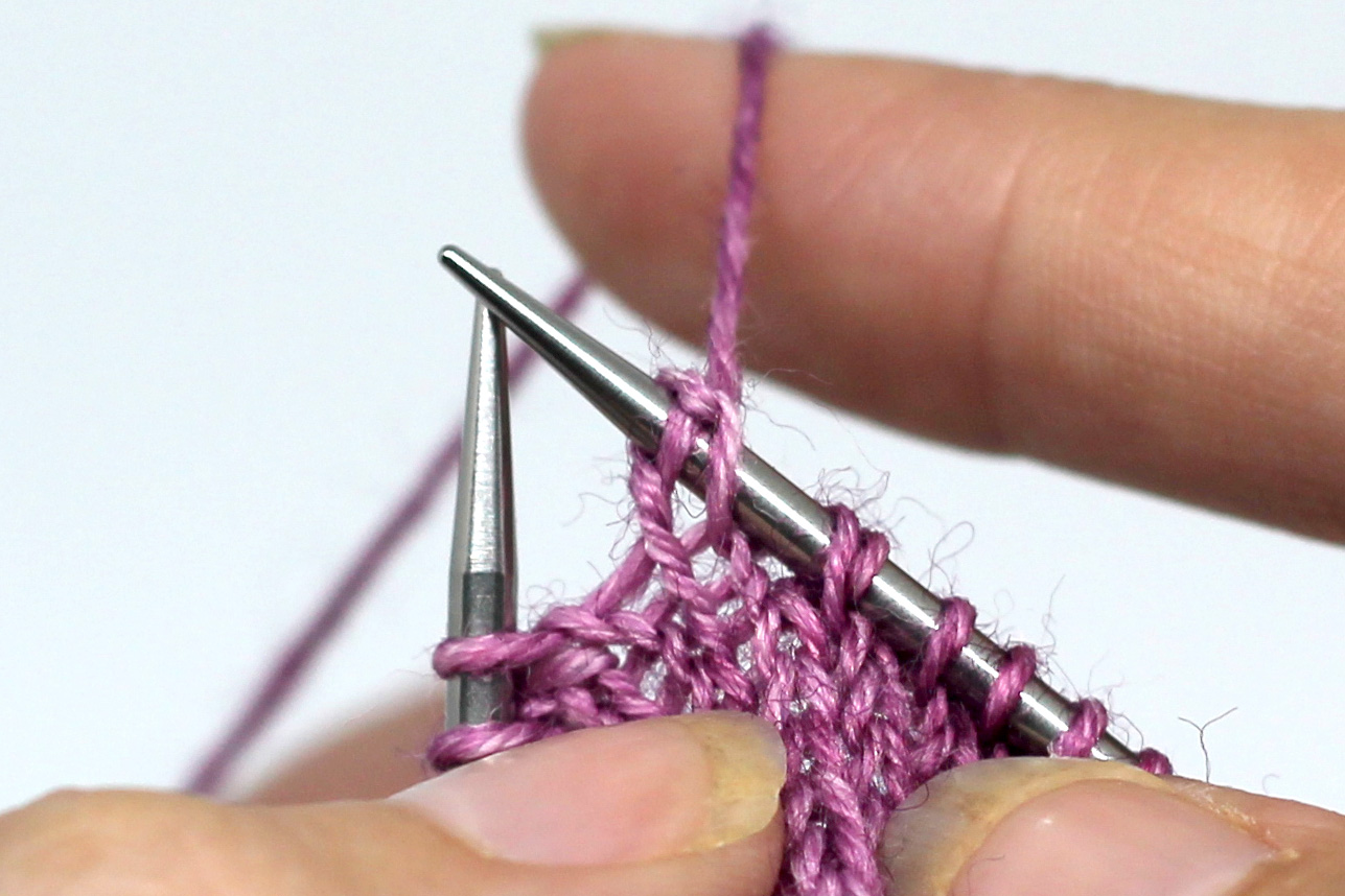 The working yarn has been pulled up over the knitting needle, causing the stitch to change shape and giving two strands over the needle instead of one.