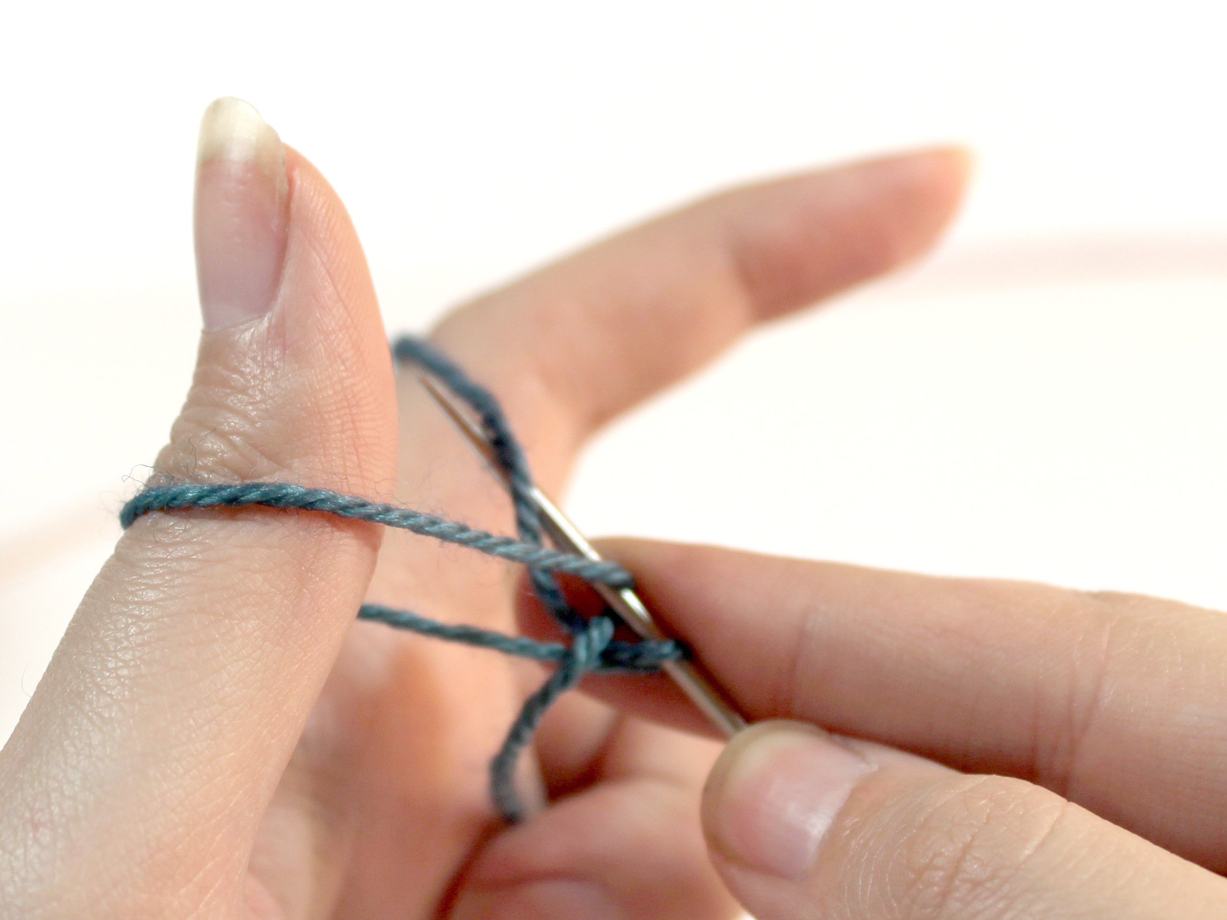 The knitting needle now has two strands of yarn from the previous steps and is catching the strand of yarn that runs from the needle to the index finger