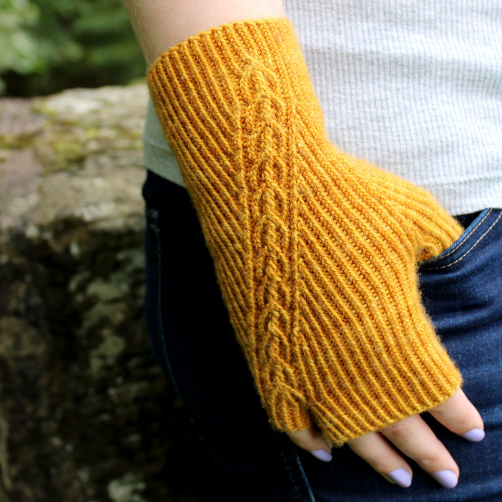 Twisted rib fingerless mitts with a narrow cable crossing the back of the hand from the inside of the wrist to the little finger. The little finger has a separate opening. Thumb is tucked into the jeans pocket of the model