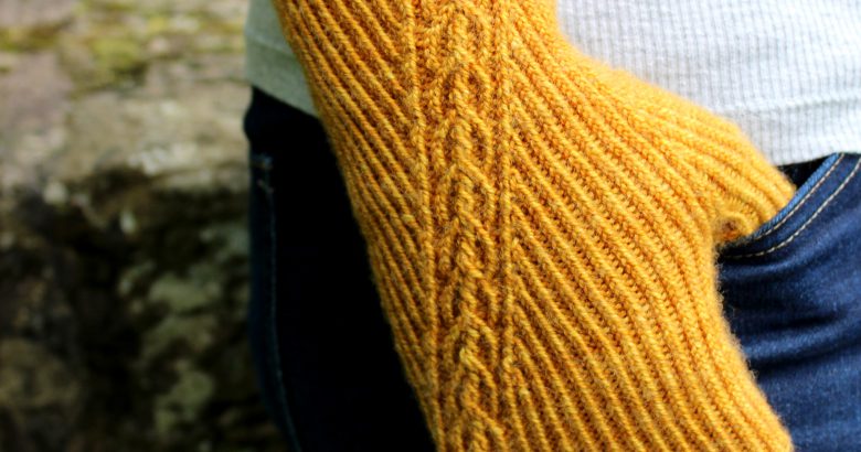 Twisted rib fingerless mitts with a narrow cable crossing the back of the hand from the inside of the wrist to the little finger. The little finger has a separate opening. Thumb is tucked into the jeans pocket of the model