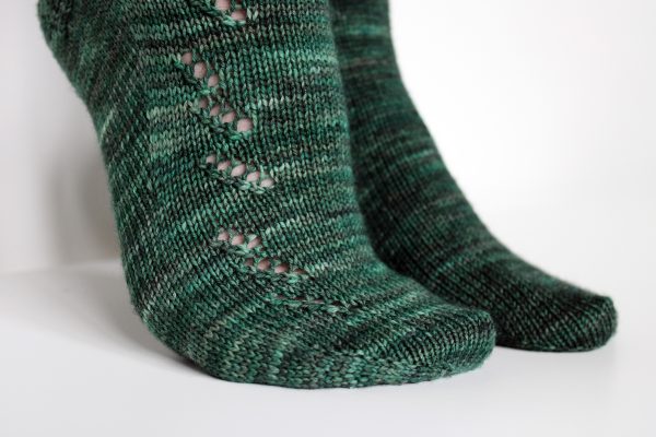 A close up on the foot of a modelled pair of socks in dark green with a lace pattern representing half a fir tree on the foot