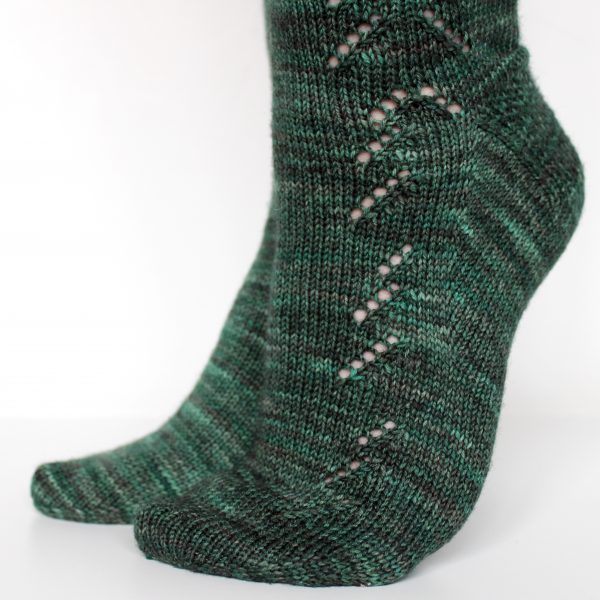 A close up on the foot of a modelled pair of socks in dark green with a lace pattern representing half a fir tree on the foot and a whole fir tree on the leg