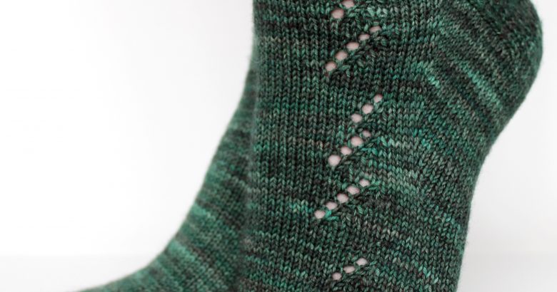 A close up on the foot of a modelled pair of socks in dark green with a lace pattern representing half a fir tree on the foot and a whole fir tree on the leg