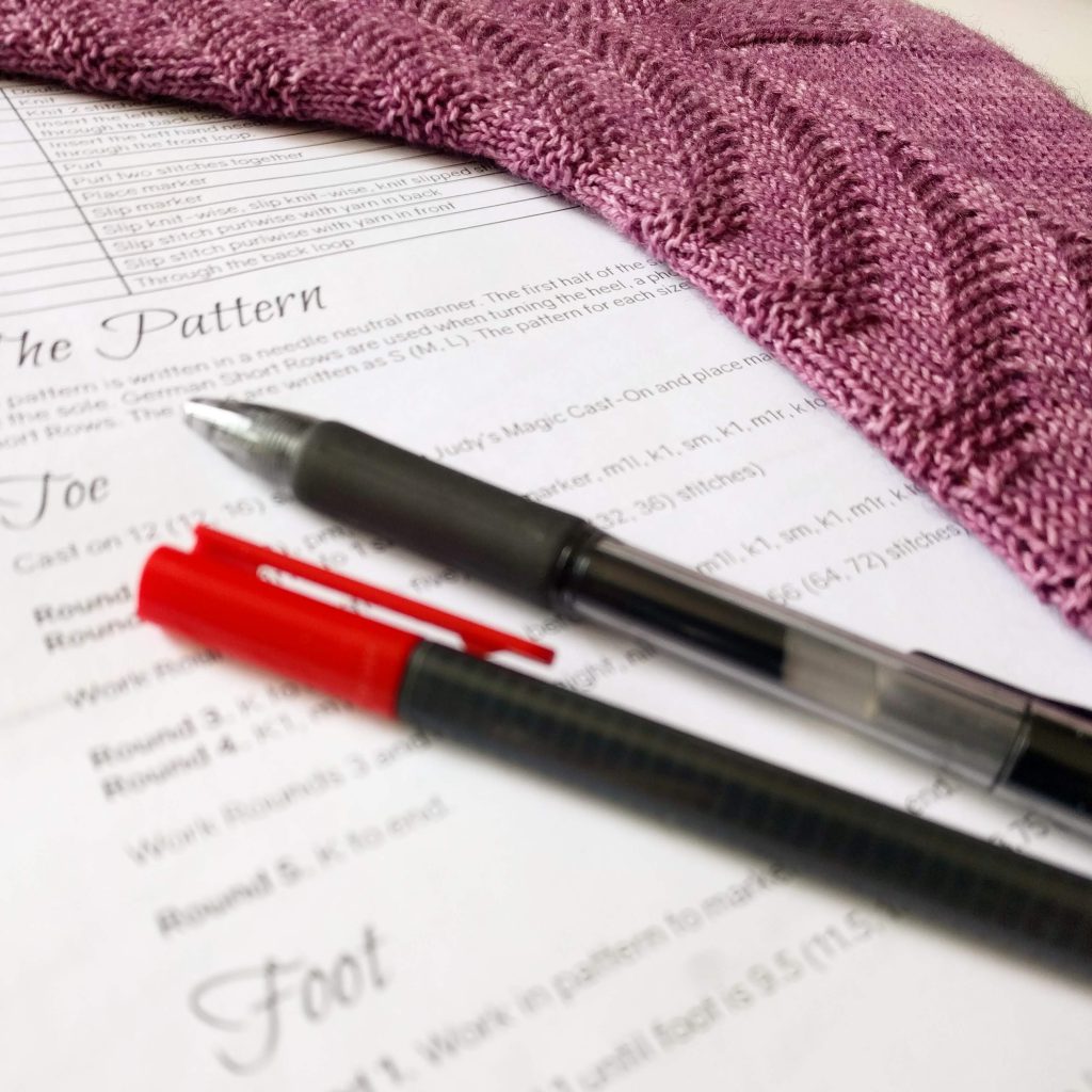 An out of focus knitting pattern with a red pen, black pen, and the sock the pattern was written for.