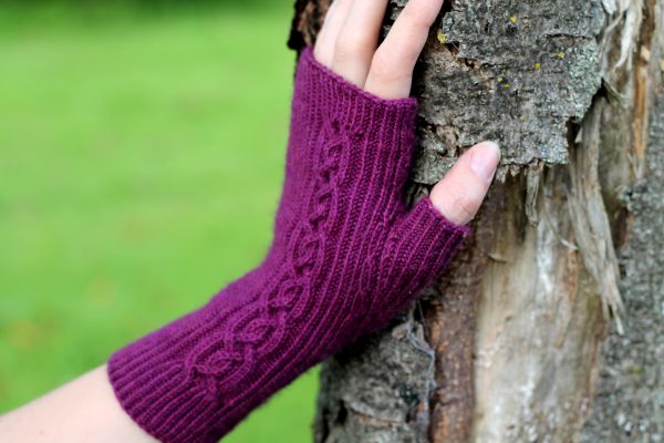 Twisted rib fingerless mitts with a large cable up the back of the hand and a narrow cable pattern that splits around the thumb (against a tree)