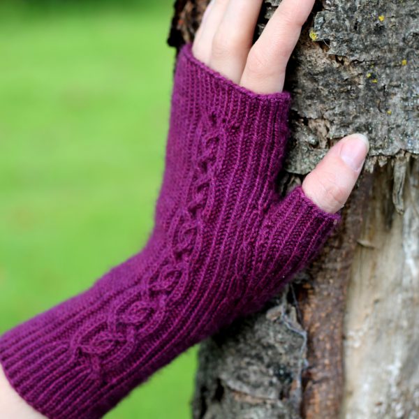 Twisted rib fingerless mitts with a large cable up the back of the hand and a narrow cable pattern that splits around the thumb (against a tree)