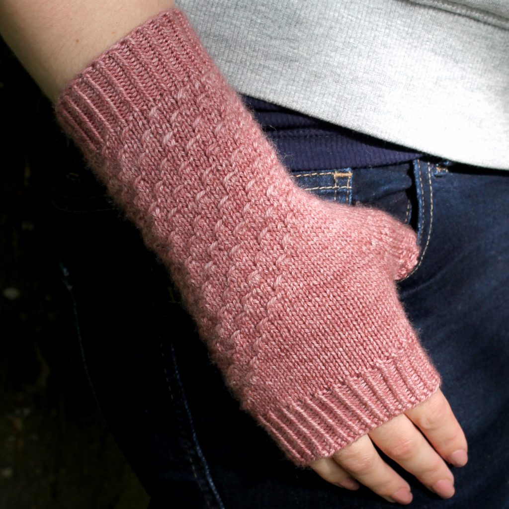 A fingerless mitt knit in pink yarn with a textured pattern travelling diagonally across the back of the hand.