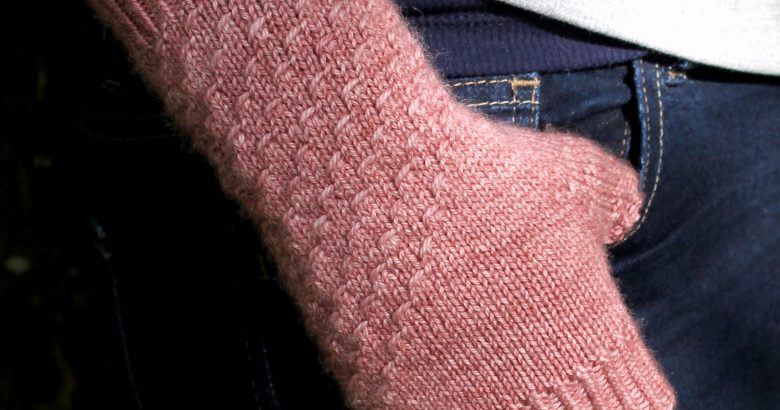 A fingerless mitt knit in pink yarn with a textured pattern travelling diagonally across the back of the hand.