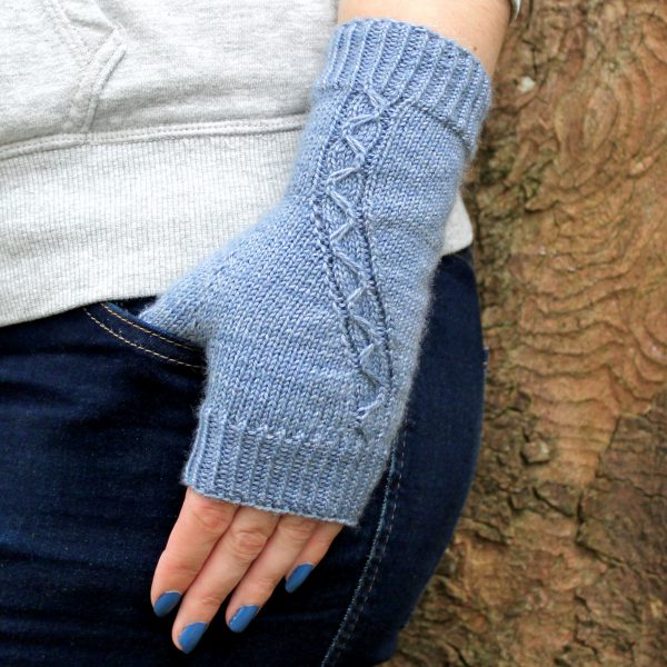 A blue fingerless mitt with a cable pattern travelling diagonally across the back of the hand, with the thumb tucked into the pocket of a pair of jeans