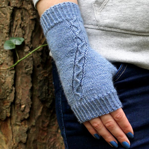 A blue fingerless mitt with a cable pattern travelling diagonally across the back of the hand, with the thumb tucked into the pocket of a pair of jeans